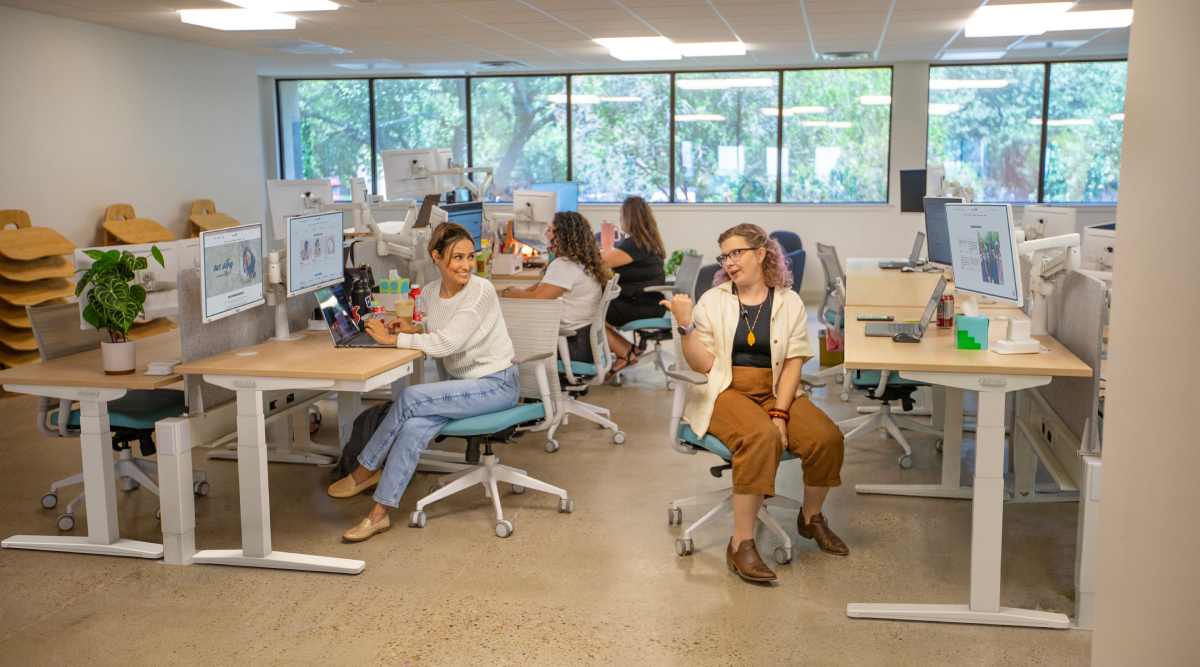 Two coworkers talking beside a height adjustable desk in an office.